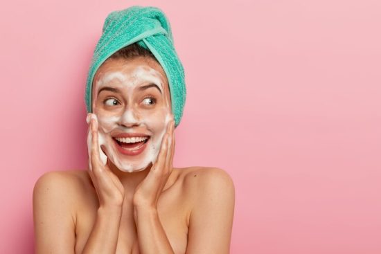 headshot-lovely-smiling-female-model-touches-cheeks-looks-aside-washes-face-with-soap-bubble-has-naked-body-wears-turquoise-soft-towel-head-poses-rosy-wall-copy-space-promotion_273609-32159